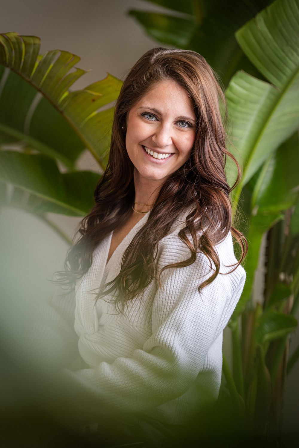 Smiling woman with long brown hair wearing a white sweater, surrounded by lush tropical green plants.