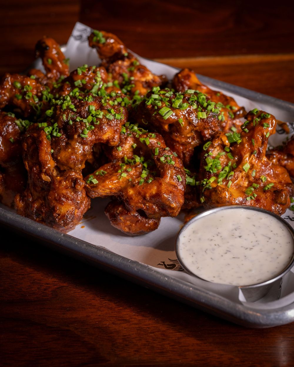 Crispy buffalo chicken wings glazed in tangy sauce, topped with fresh herbs, served with creamy ranch dip on a metal tray.