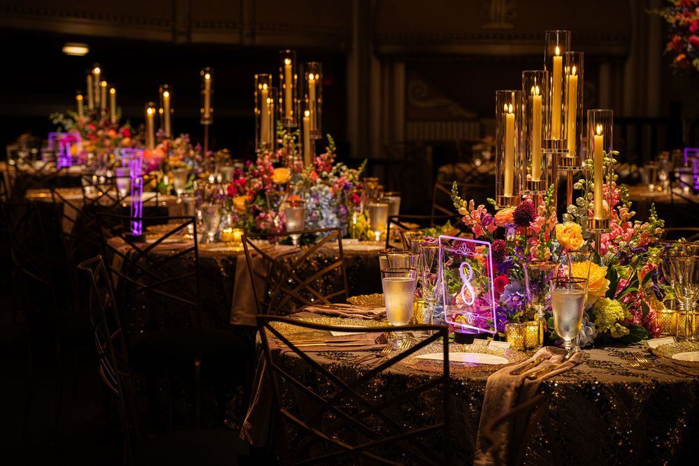 Elegant wedding reception table with gold candelabras, colorful floral arrangements, and glowing candles on a dark sequin tablecloth.