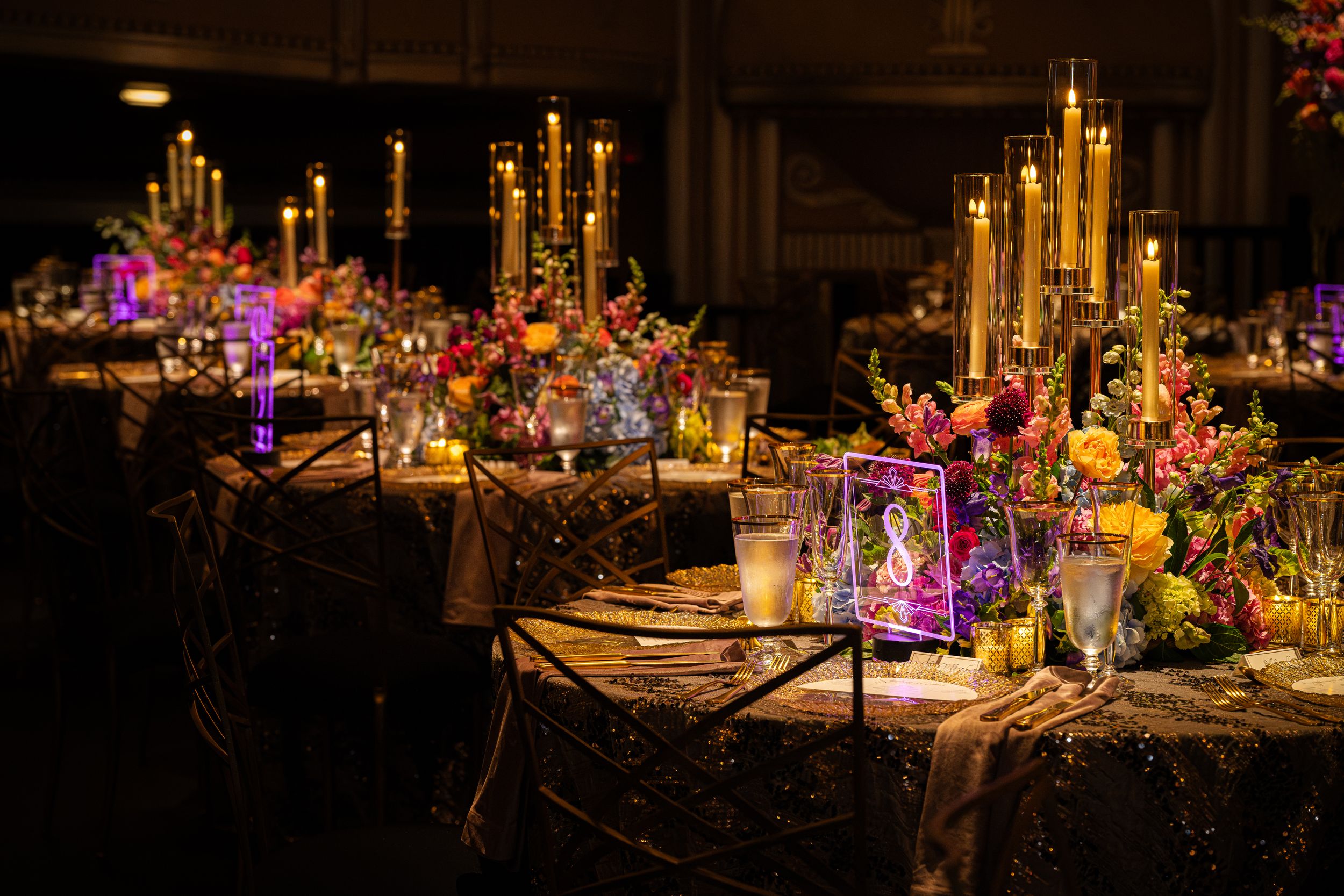 Elegant wedding reception table with gold candelabras, colorful floral arrangements, and glowing candles on a dark sequin tablecloth.