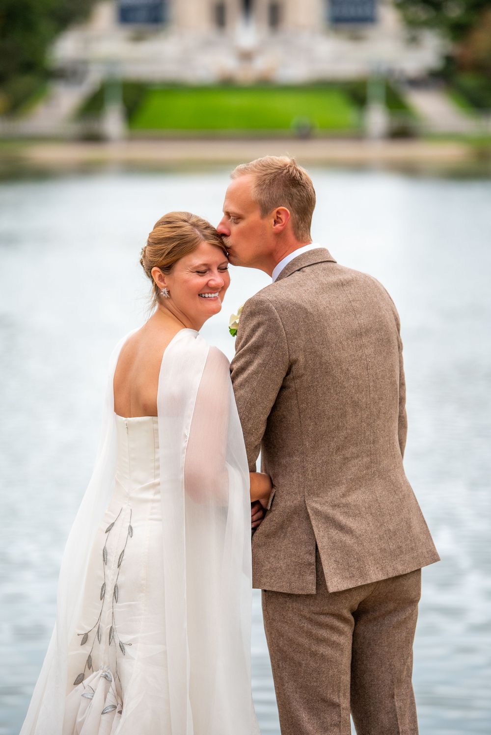 A couple shares a romantic moment by a lake with the person wearing a white dress and veil.