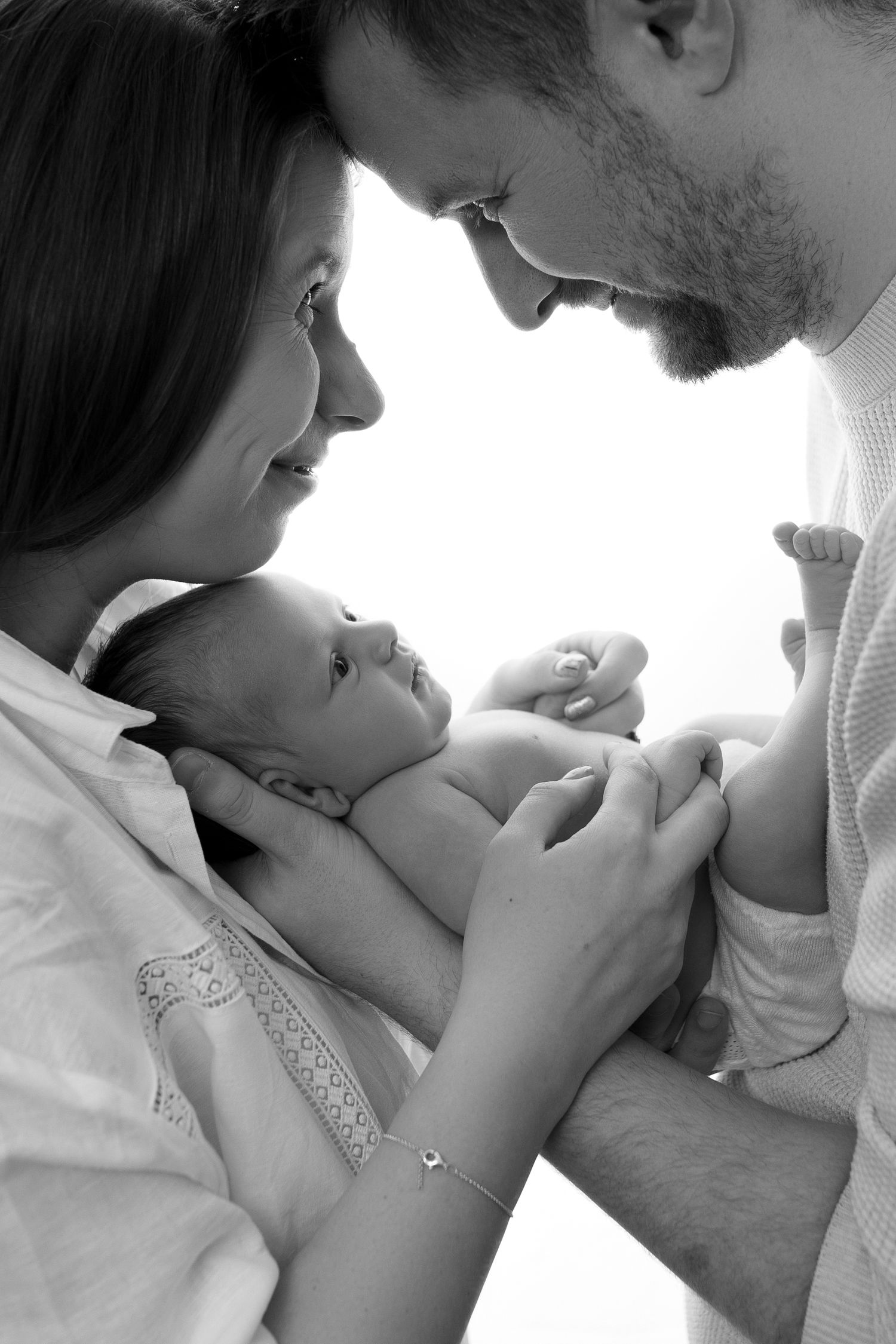 A tender black and white portrait shows a couple gazing lovingly at their newborn baby between them.