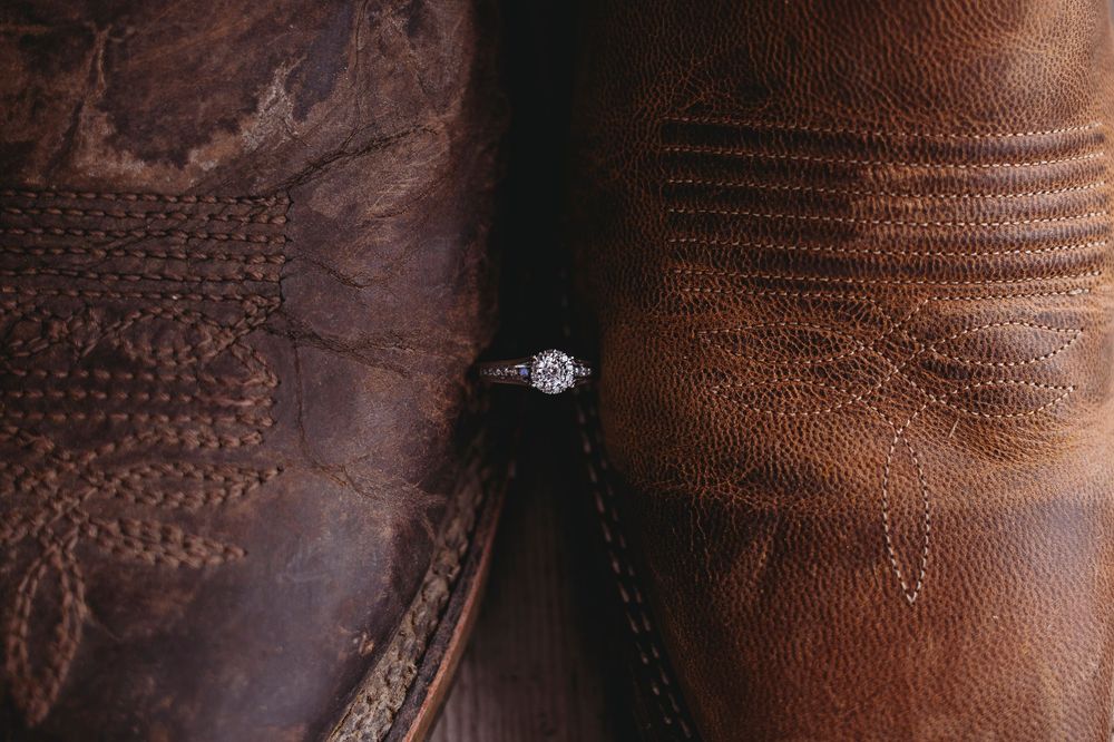 Diamond engagement ring nestled between two brown leather cowboy boots, rustic western wedding proposal photography.