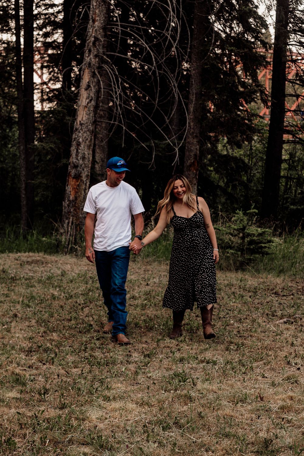 A couple holding hands while walking through a wooded area at sunset, wearing casual summer attire.