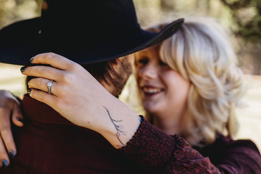 Smiling blonde woman embracing a partner in a black hat, showing a delicate wrist tattoo and ring outdoors.