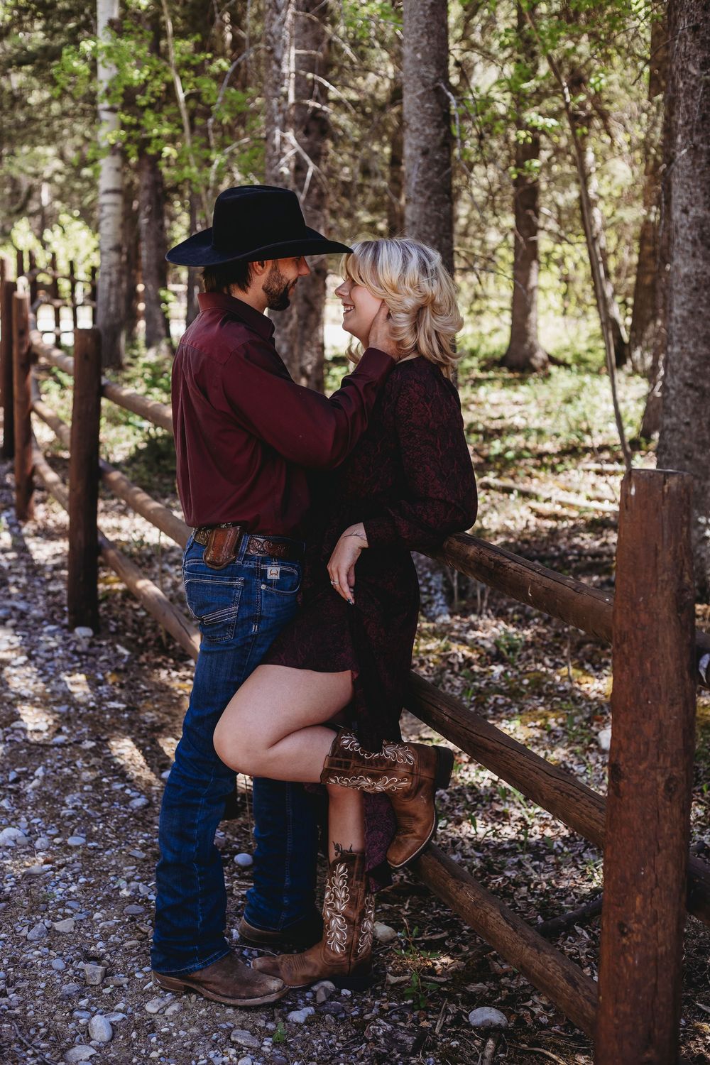 Couple in Western attire sharing a romantic moment by a rustic wooden fence in a forest setting.