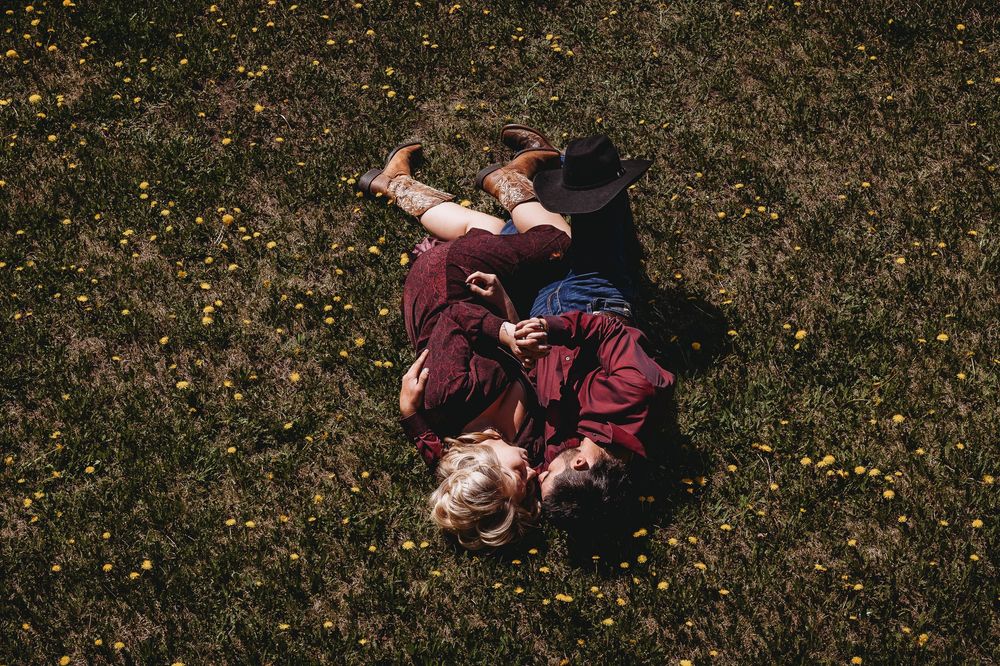Two children lying on a green grass field with yellow wildflowers, viewed from above on a sunny day.
