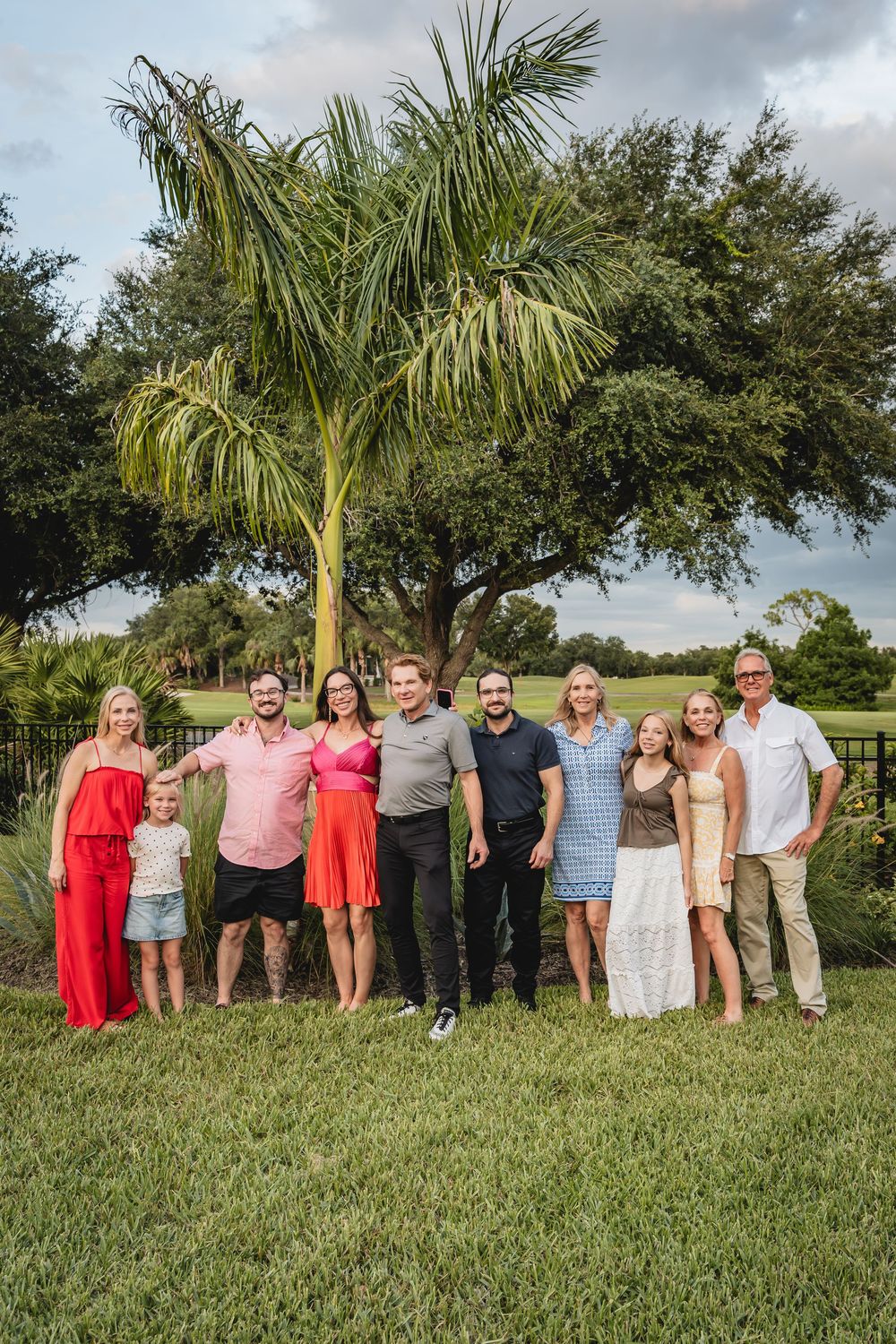 Large family group of nine people smiling together on a green lawn with palm and oak trees in background. Family Photo #familyphotographer