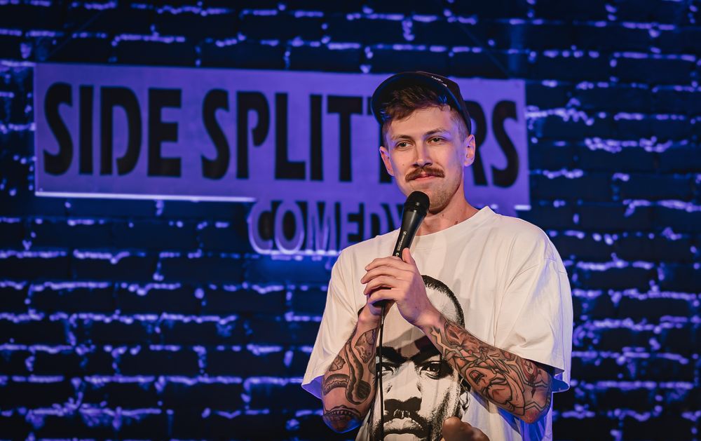 A tattooed comedian performs stand-up on stage at Side Splitters Comedy Club holding a microphone. Cam Bertrand