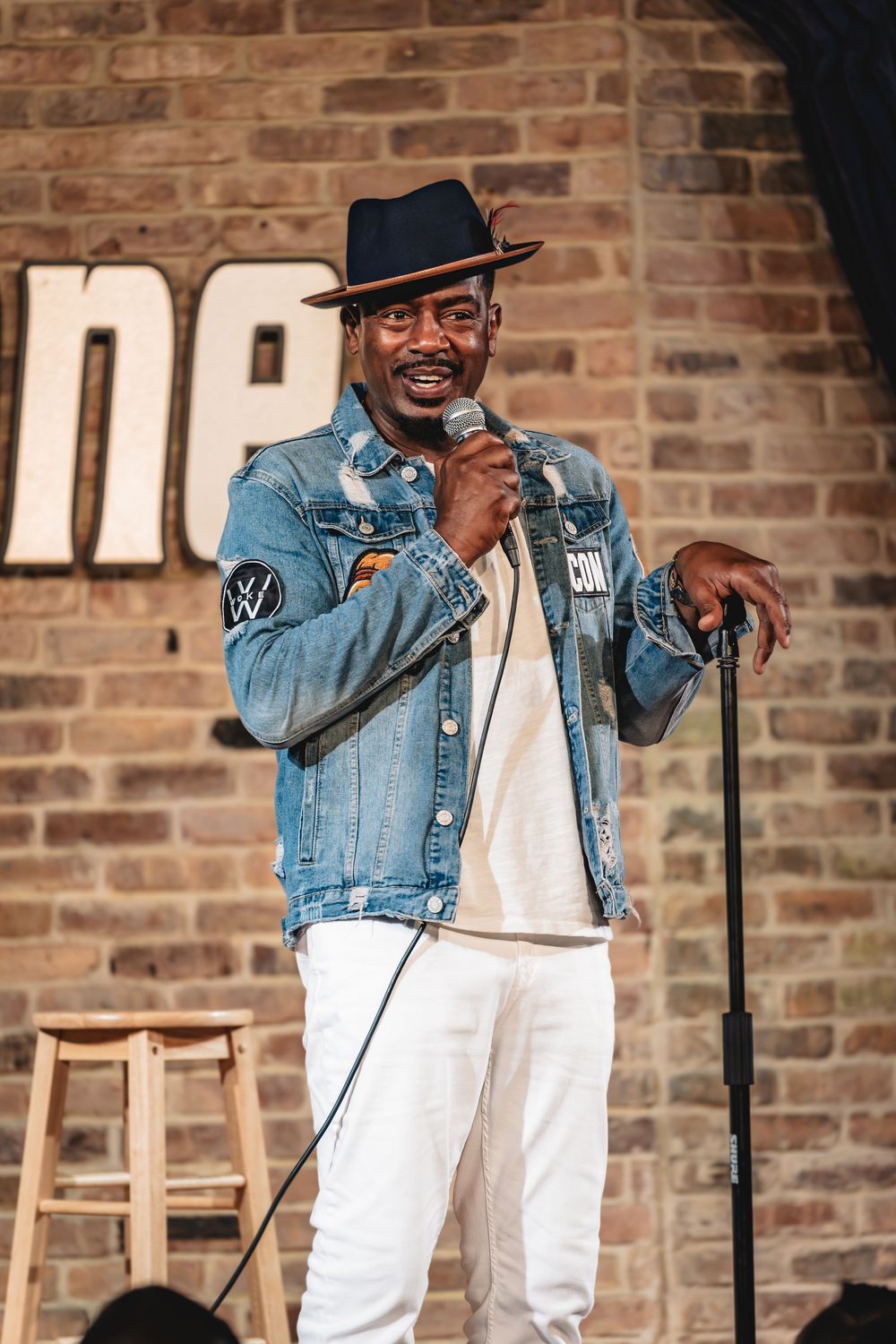 Bill Bellamy, Stand-up comedian performing on stage wearing denim jacket, white pants, and black hat, holding microphone at comedy club.