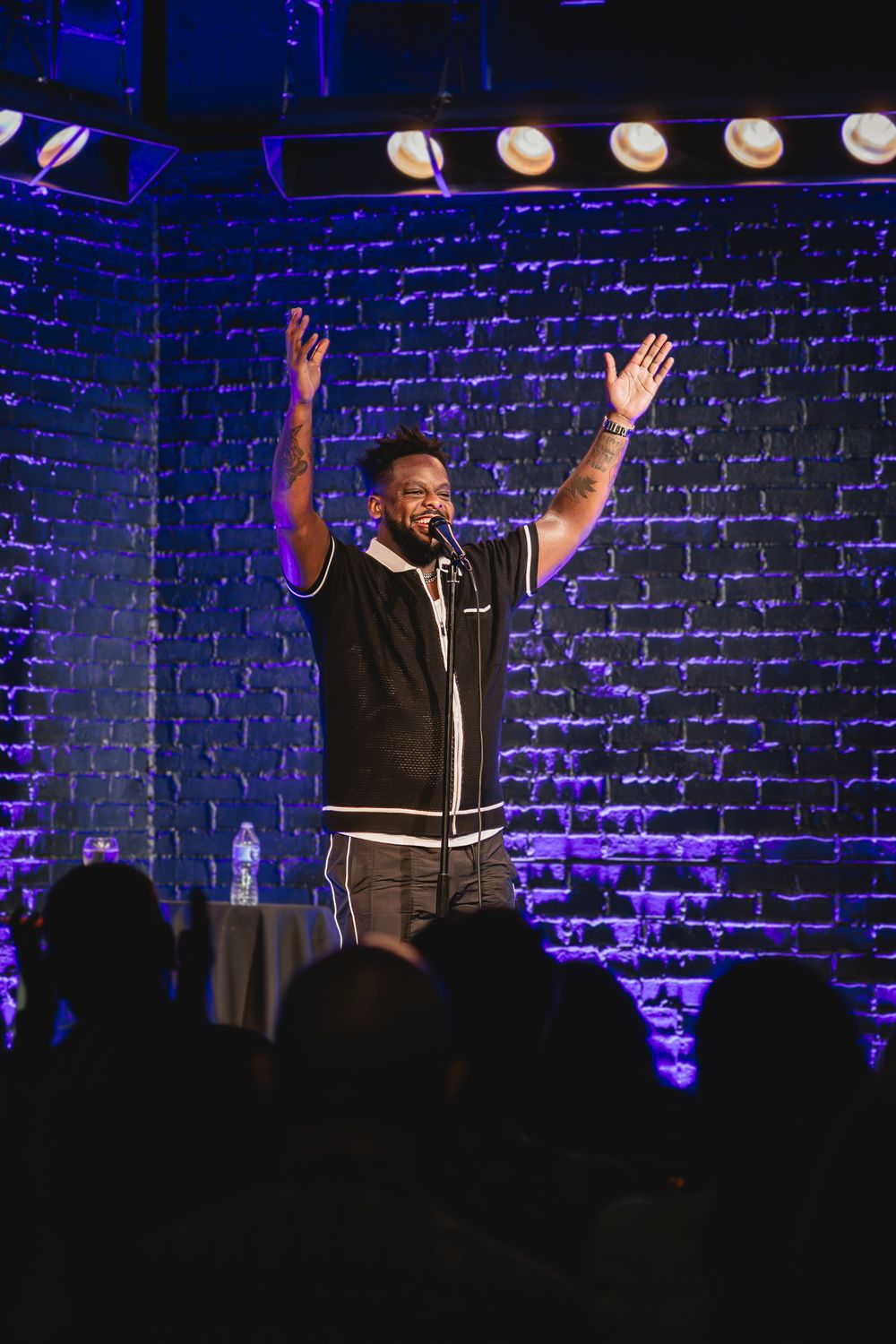 Comedian performs on stage with hands raised at a brick-walled comedy club with purple lighting. Dedrick Flynn at Side Splitters Comedy Club