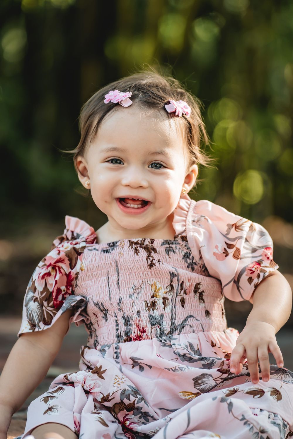 Smiling baby girl in a floral pink dress with hair clips, sitting outdoors in a sunlit garden setting.