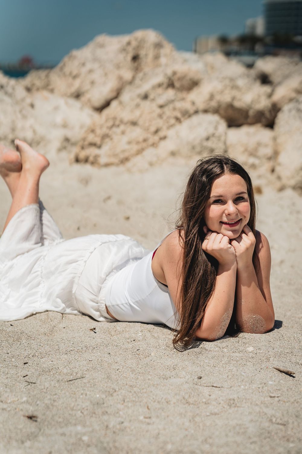 Young woman in white dress lying on sandy beach, smiling with hands under chin, rocky backdrop on sunny day. Graduation photos, senior photo