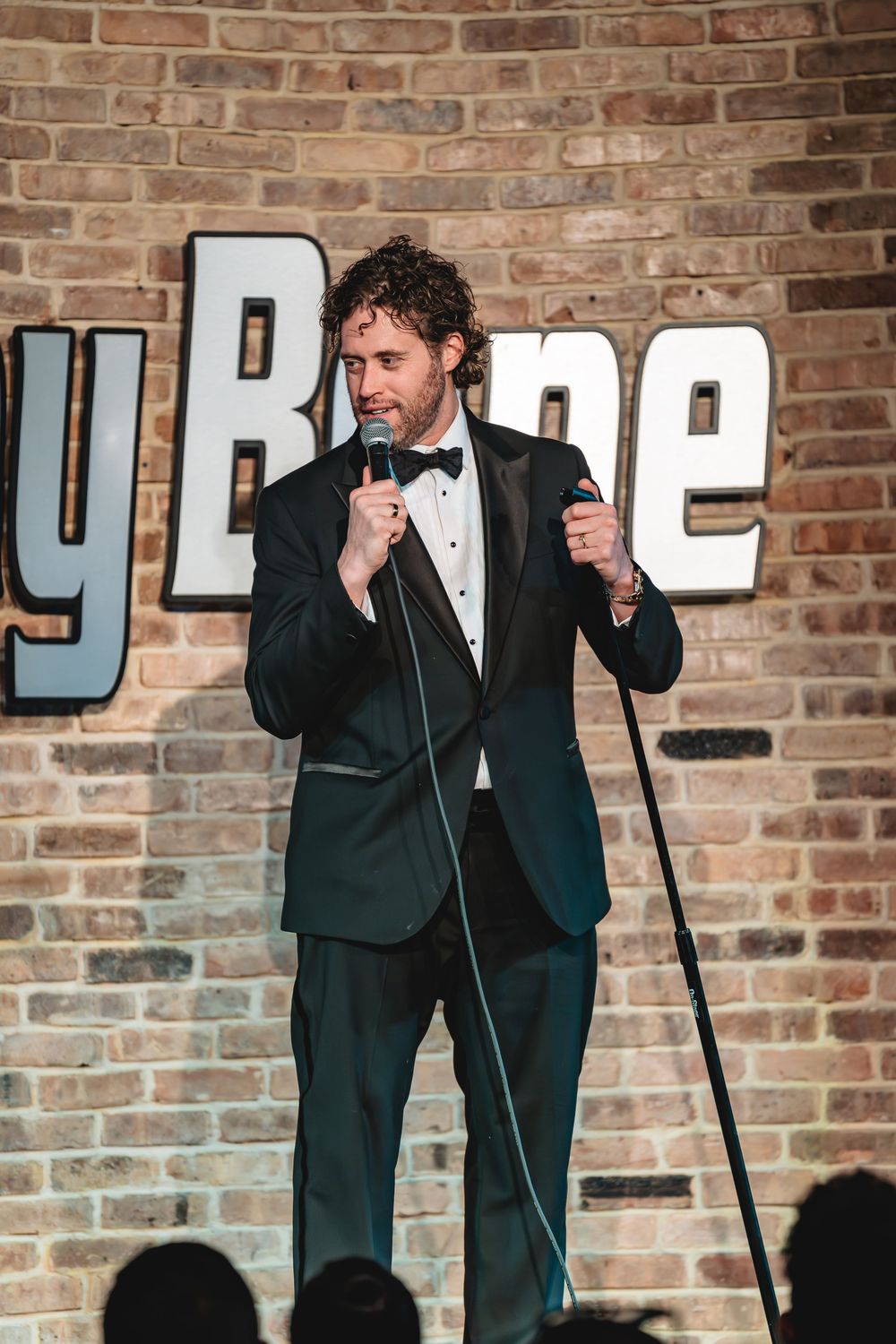 Comedian in tuxedo performs stand-up at Comedy Bone club, holding microphone on brick wall stage. T J Miller, comedy photos, comedy video