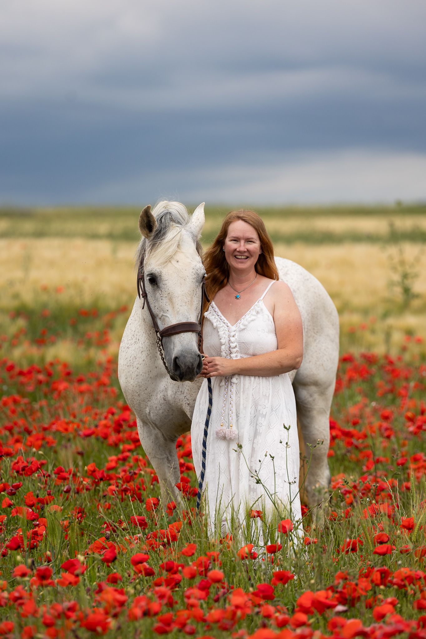 Woman in white dress standing with a white horse in a vibrant red poppy field under a dramatic cloudy sky.
