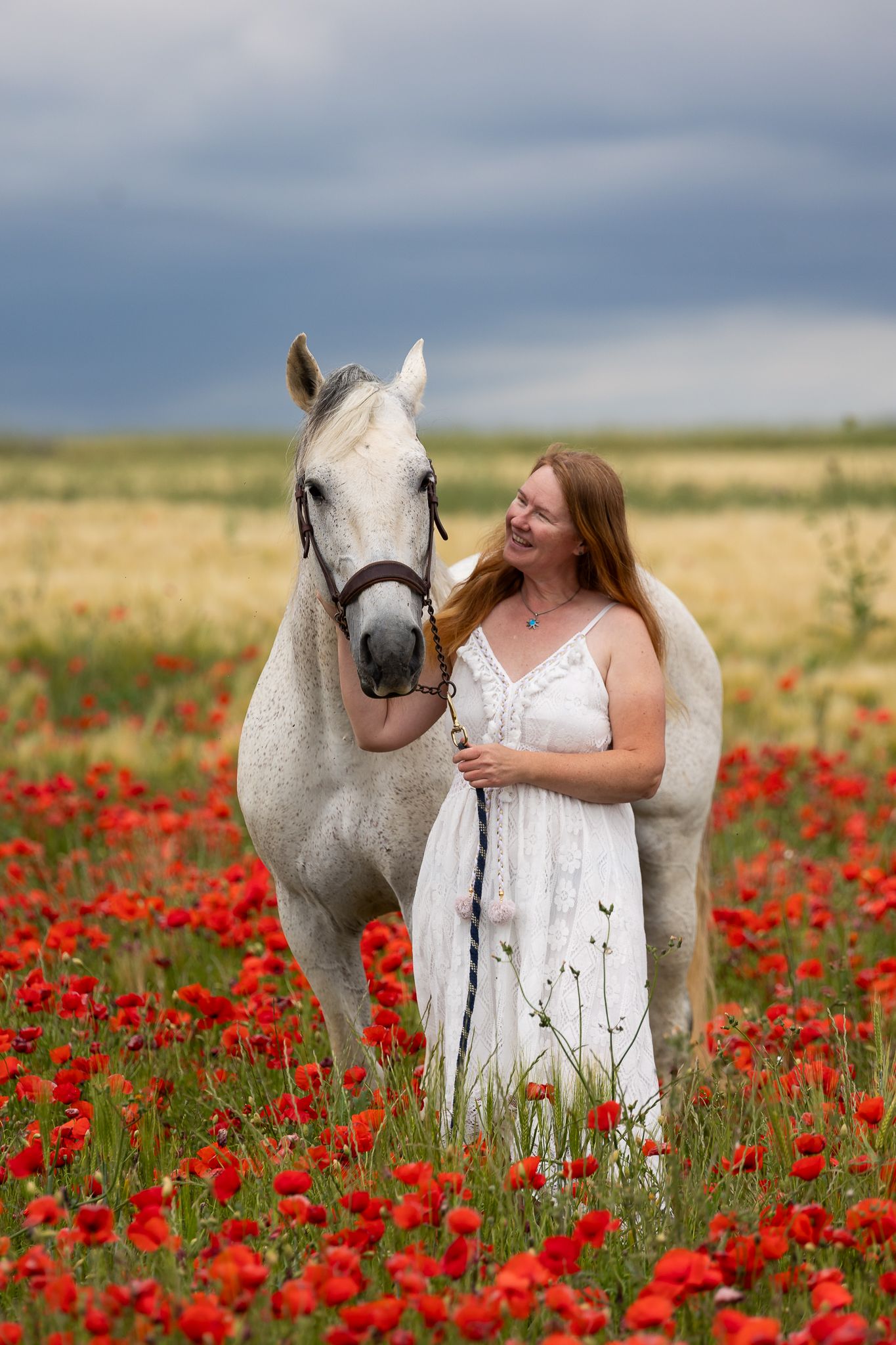 Woman in white dress smiling beside a white horse in a vibrant red poppy field under a cloudy sky.