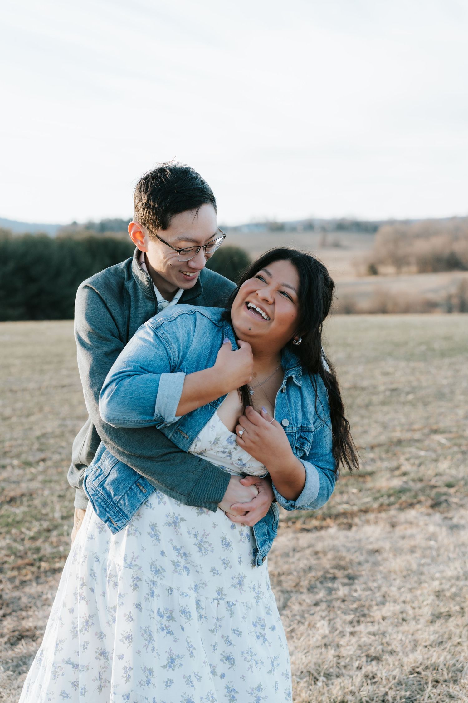 A joyful couple embraces and laughs together in a field during golden hour while wearing denim jackets.
