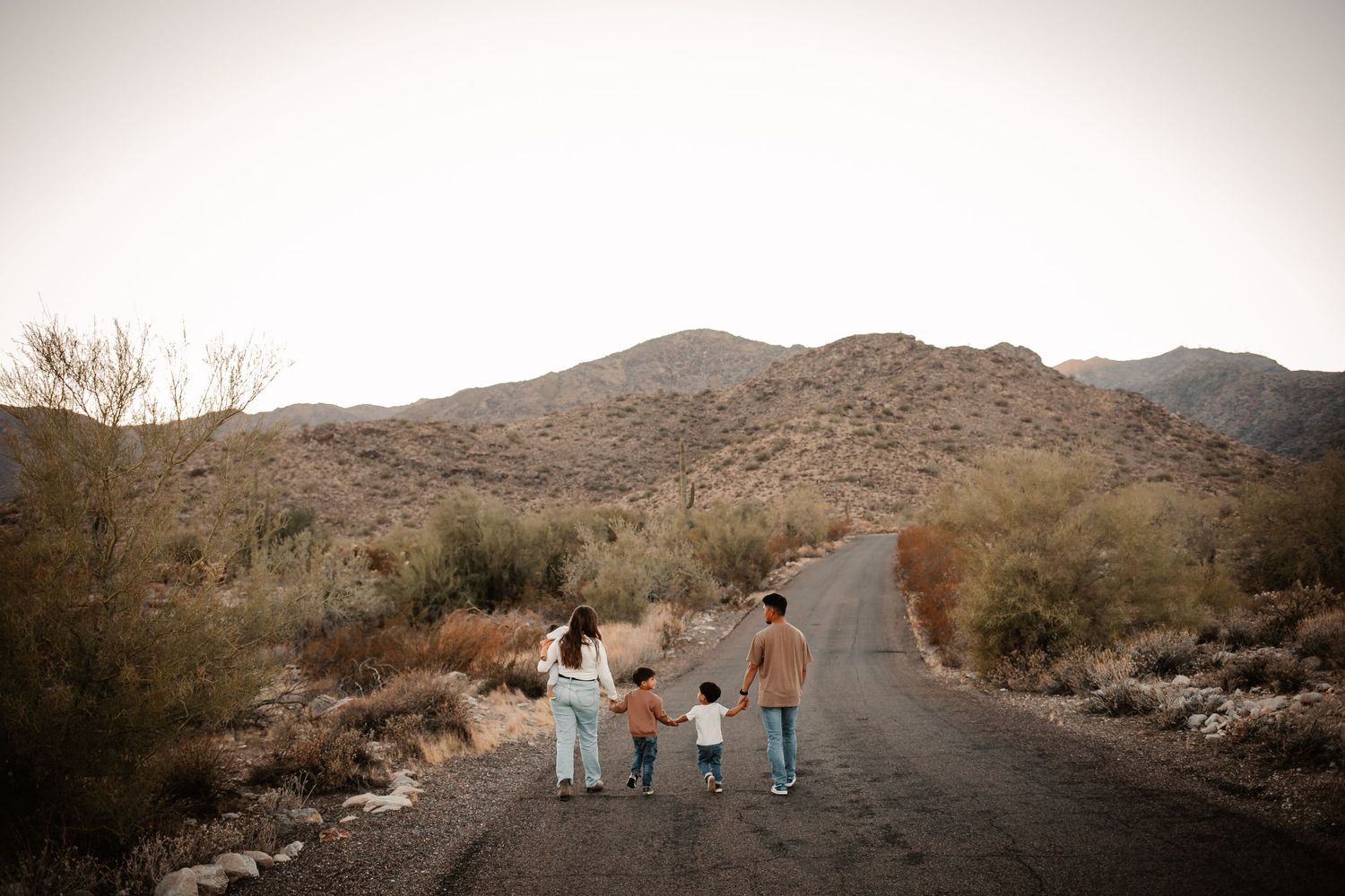 Adorable toddler standing between two adults in jeans and sneakers on a sandy outdoor path.