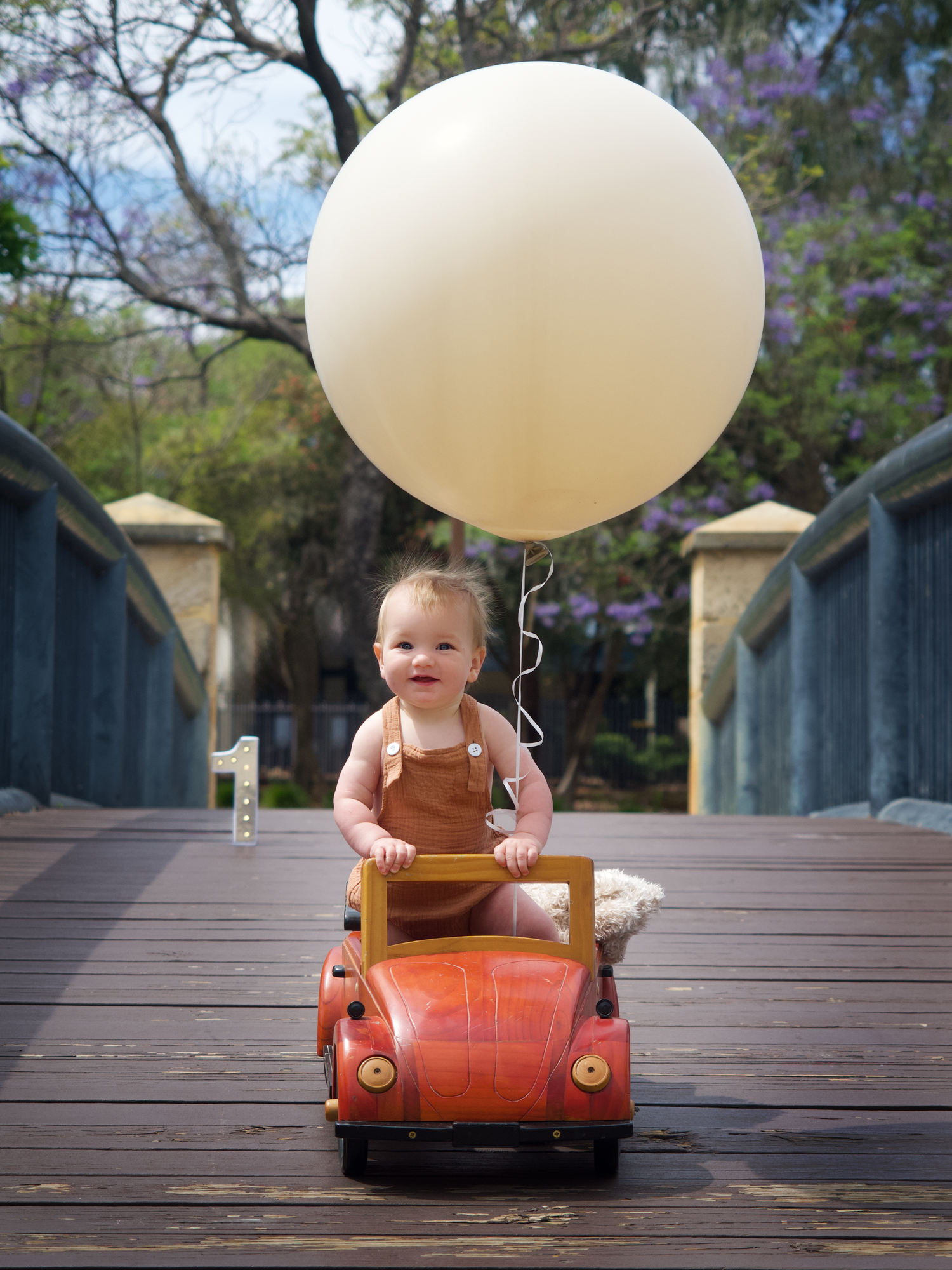 baby boy first birthday standing in a wooden car holding a balloon on a bridge