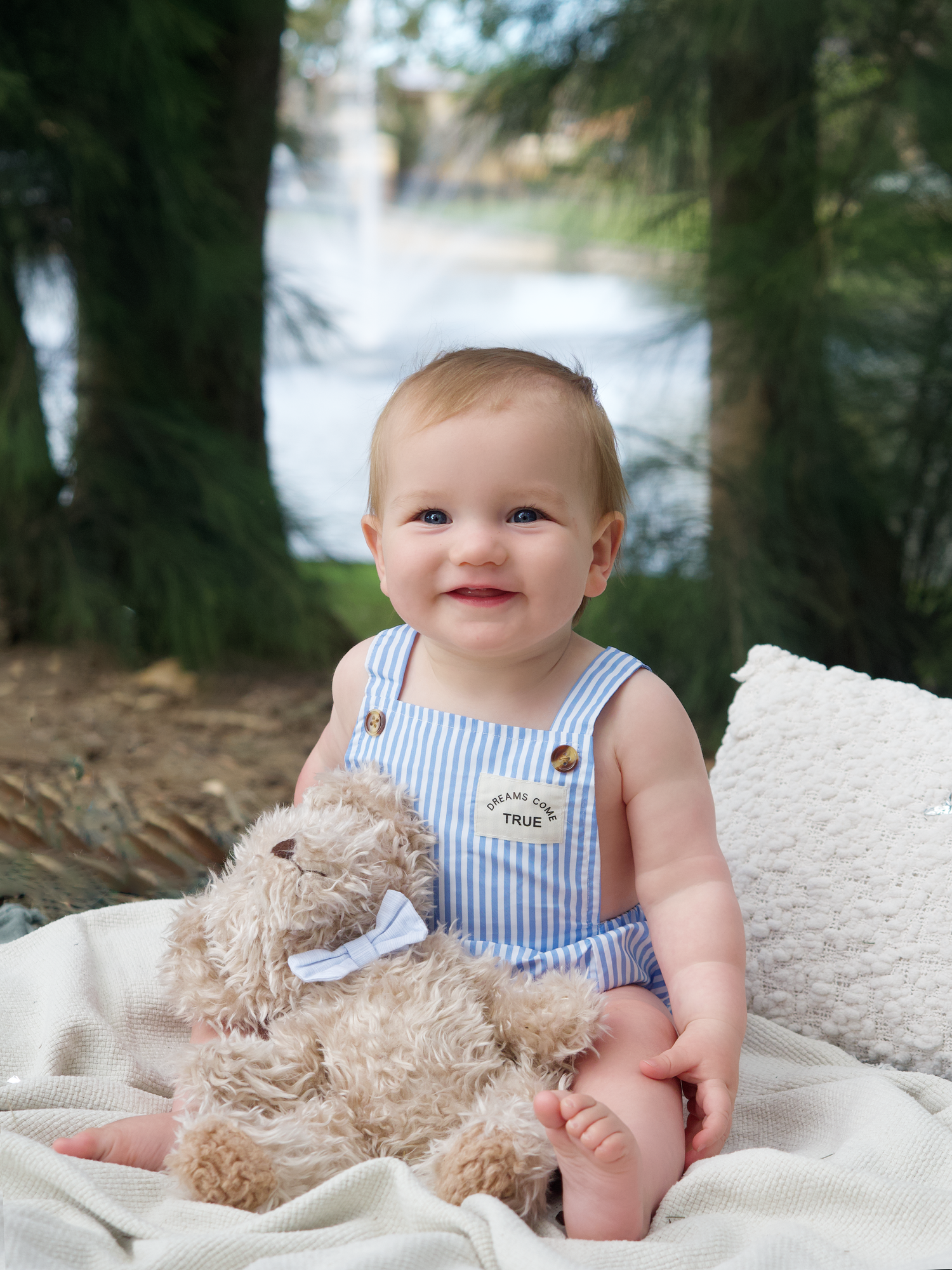 outdoor photo session of a boy first birthday smiling sitting on a rug with a teddy