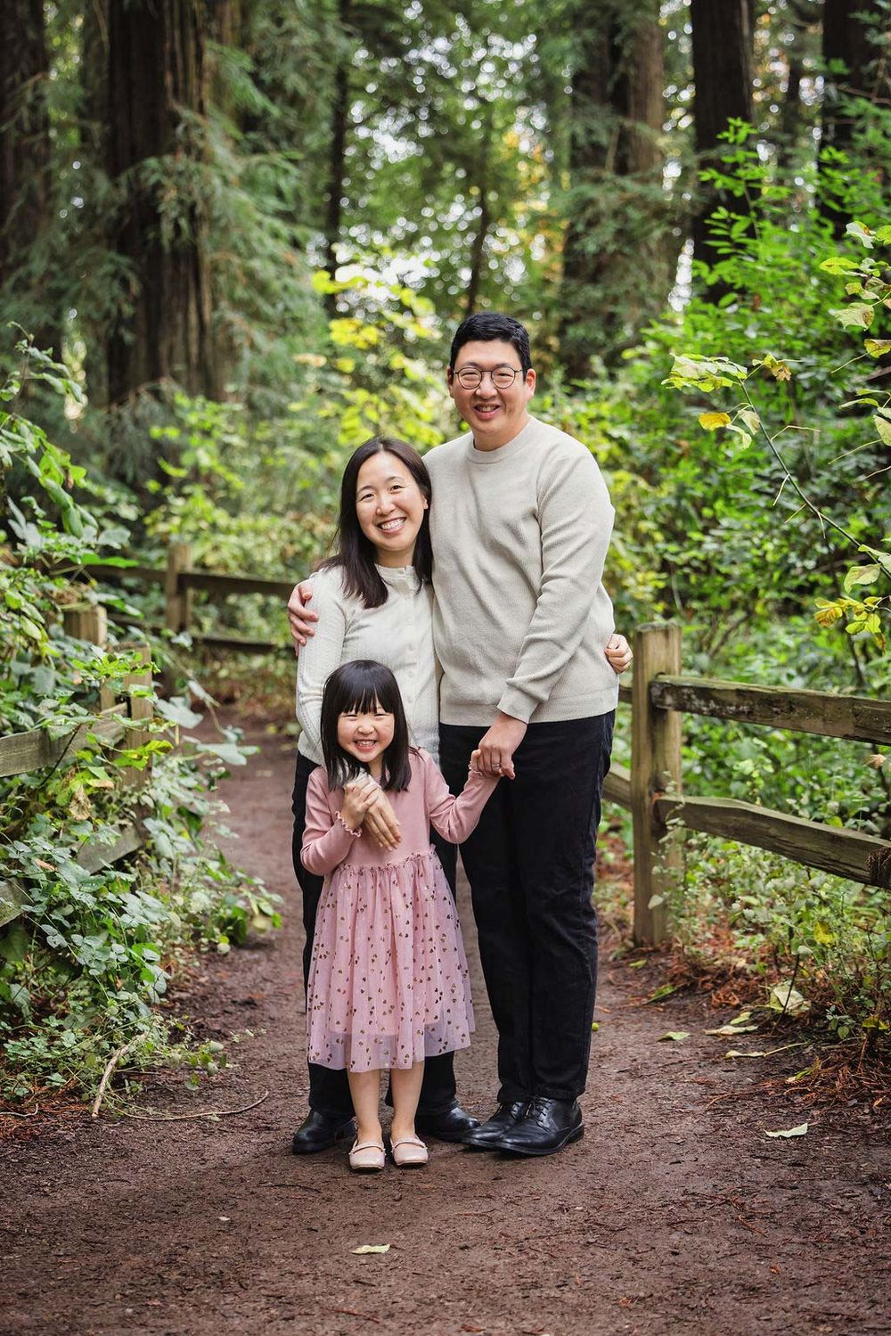 Asian family of three smiling on a forest trail, parents with young daughter in pink dress holding hands.