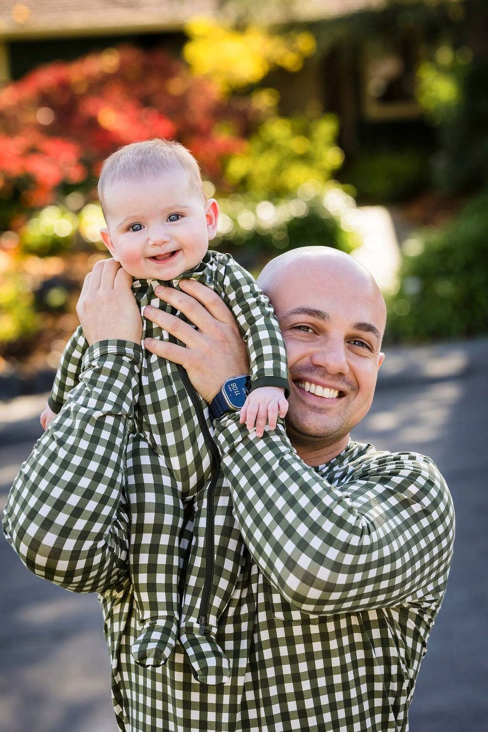 Smiling bald father holding matching outfit baby boy outdoors with colorful foliage background.