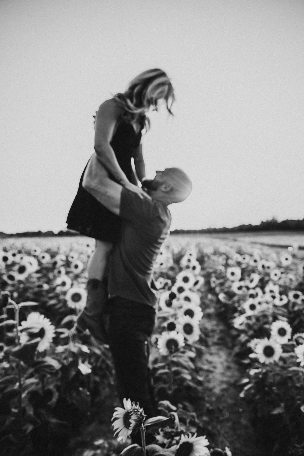 Black and white image of someone standing in a sunflower field with their face obscured against a cloudy sky.