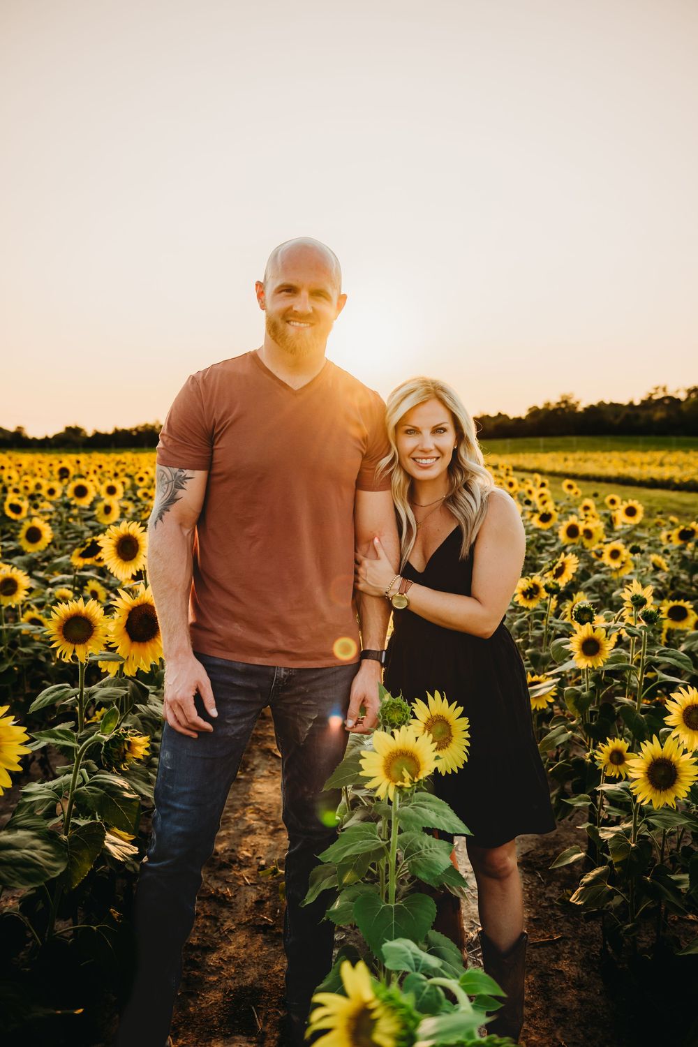 A couple stands together in a beautiful sunflower field at sunset with golden light streaming through the flowers.
