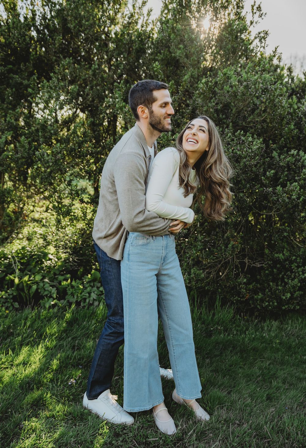 A couple embraces and laughs together outdoors on a sunny day with greenery in the background.