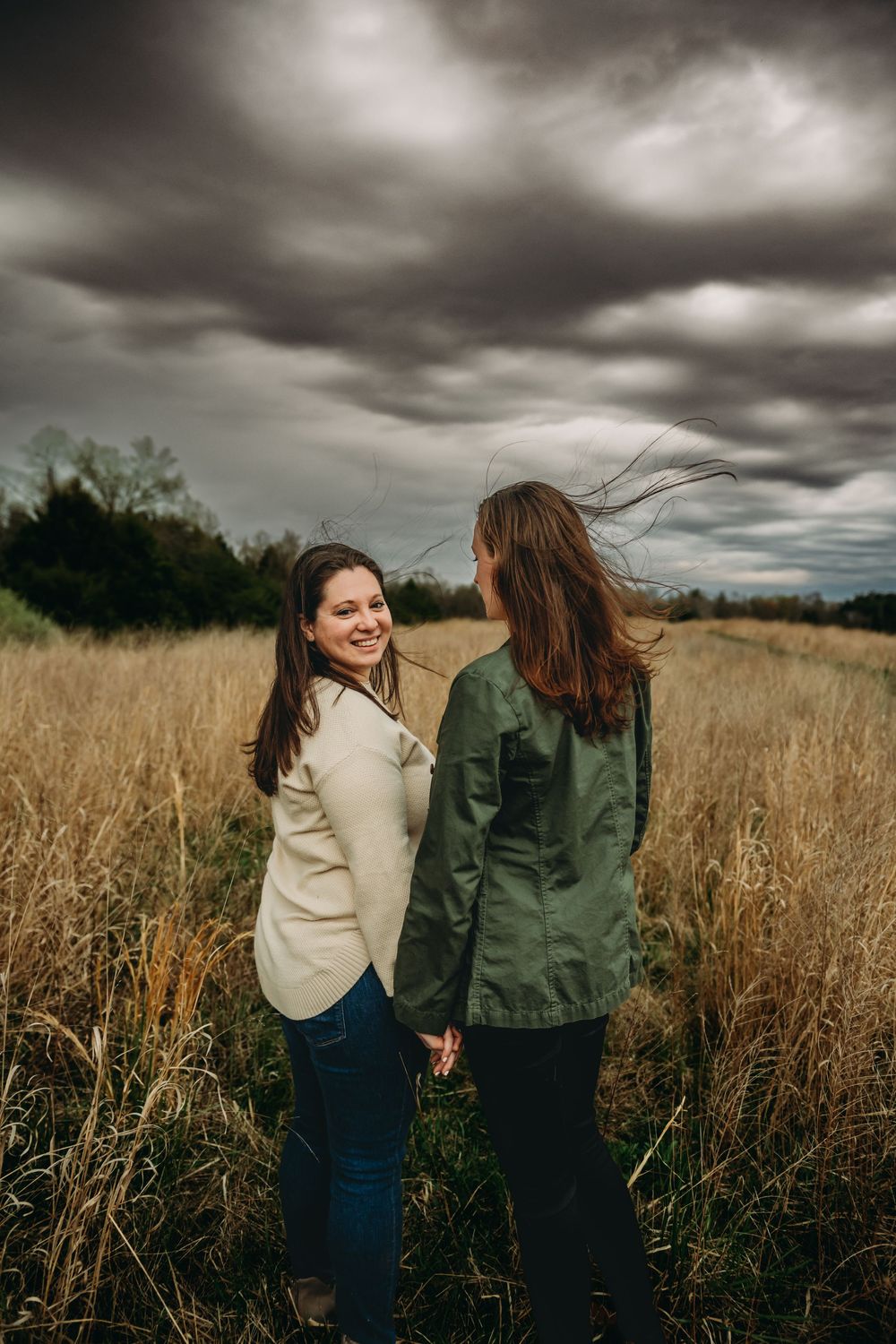 Two friends share a candid moment in a tall grass field under dramatic stormy clouds.
