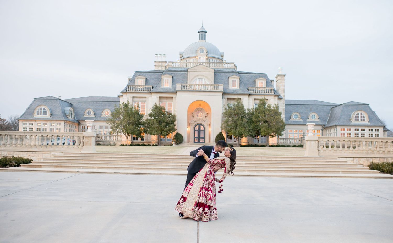 Couple in traditional Indian wedding attire share a romantic dip pose in front of a grand, elegant château-style estate. The Olana Texas