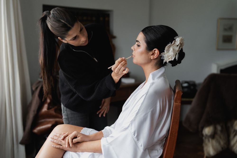 A makeup artist applies makeup to a bride in a white satin robe with a floral hair accessory while she sits during wedding preparations.