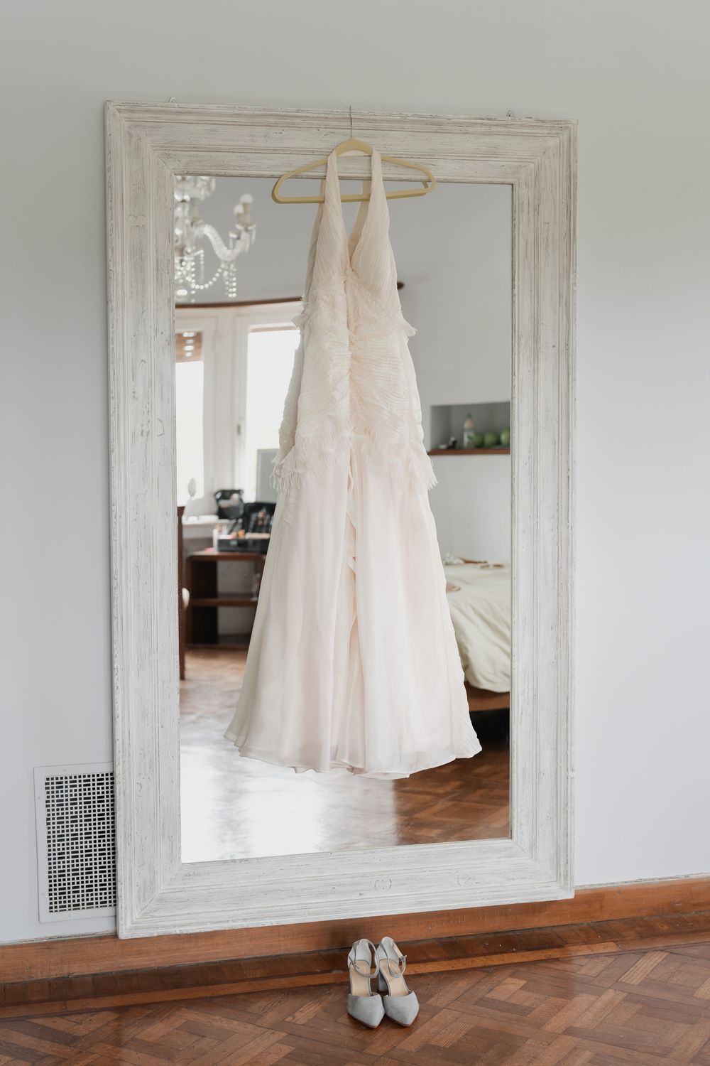 Ivory wedding dress hanging on a wooden hanger reflected in a large white-framed mirror, with bridal shoes on the floor below.