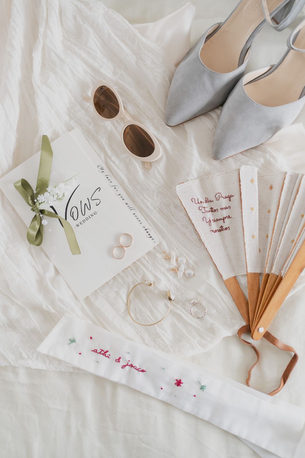 Flat lay of wedding vows booklet, sunglasses, gold rings, silver heels and a hand fan on white linen.