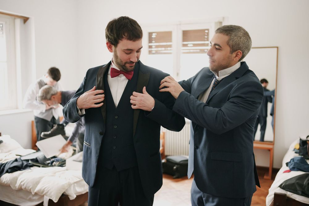 A groom in a vest and red bow tie gets help adjusting his suit jacket from a groomsman in a bright, sunlit room before the wedding.