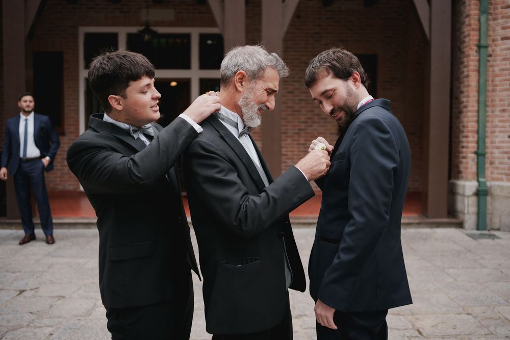 Three men in dark suits help each other adjust a boutonniere and lapel outside a brick building before a wedding ceremony.