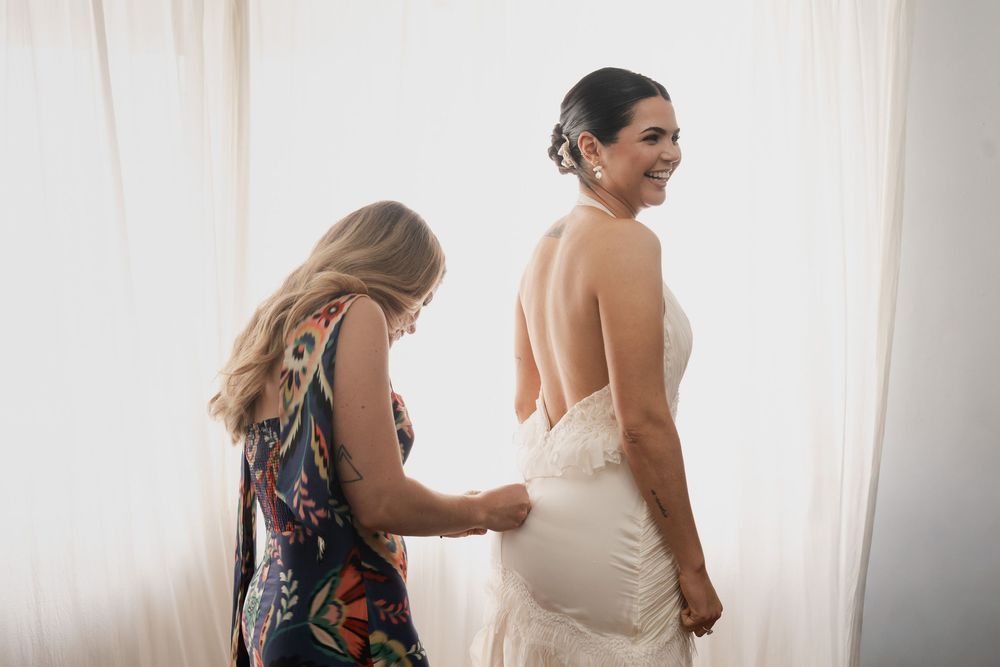 A smiling bride in an elegant backless white wedding gown gets help fastening her dress from a woman in a colorful patterned outfit.