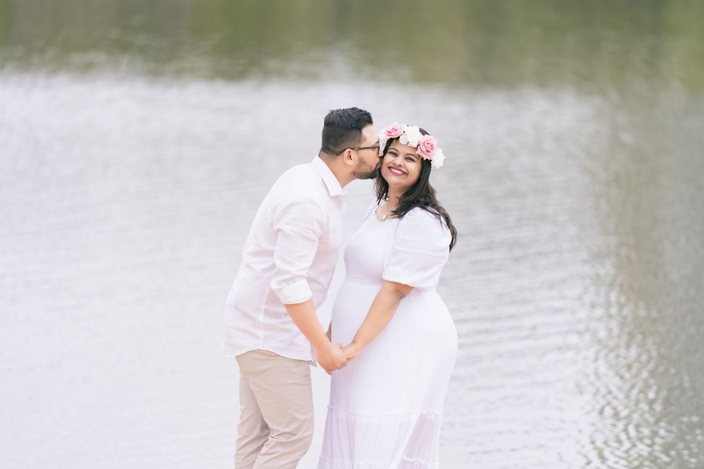 A man in a white shirt kisses the cheek of a smiling woman in a white dress and pink flower crown while standing by a calm lake.