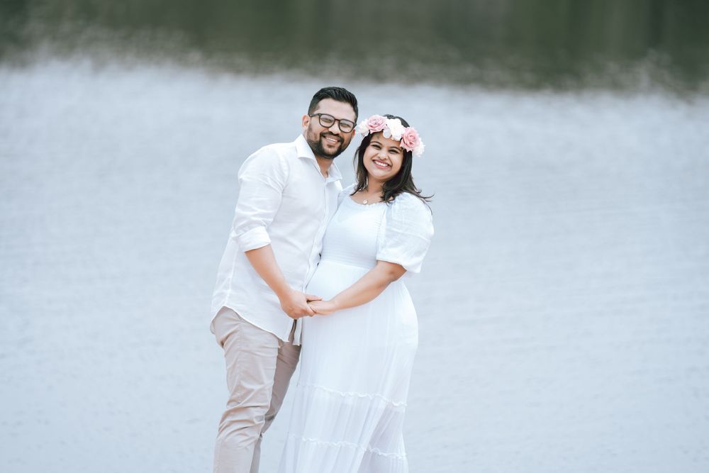 Smiling couple in white outfits holding hands by a calm lake during an outdoor maternity photoshoot, the woman wearing a pink flower crown.