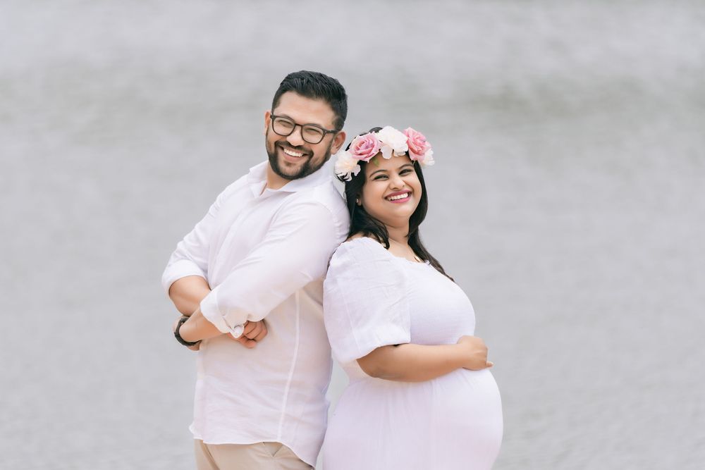 Smiling couple standing back to back in white outfits during a maternity photoshoot, the pregnant woman wearing a pink floral flower crown.