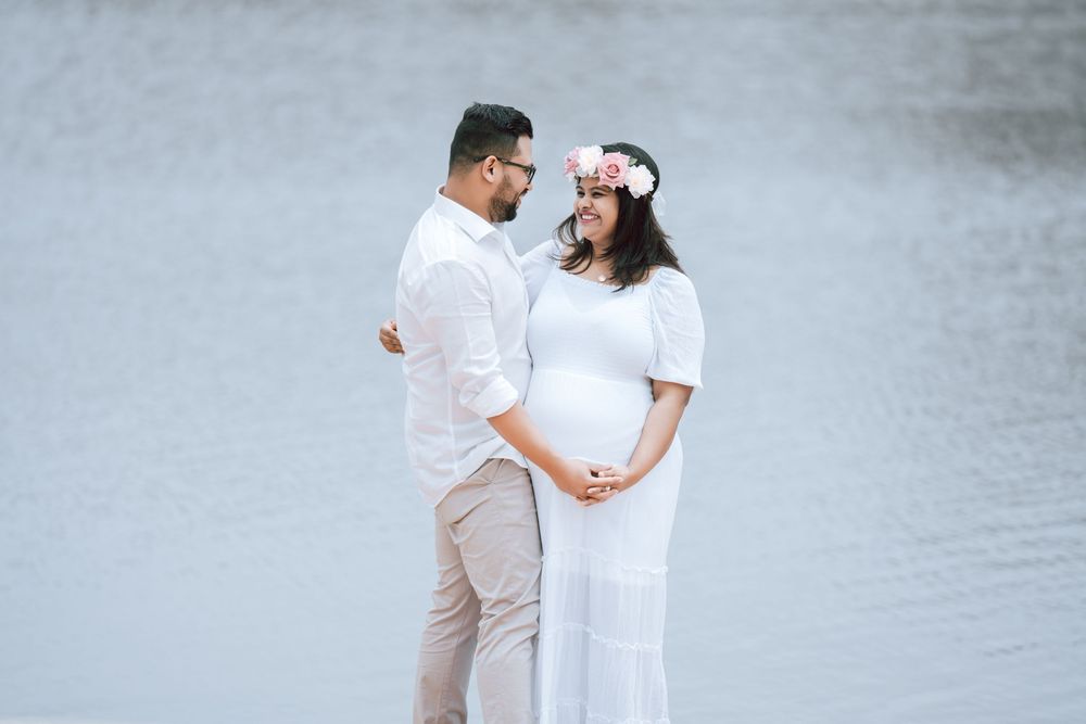 Expecting couple in white outfits embrace and hold the baby bump during a maternity photo shoot beside calm water, she wears a floral crown.
