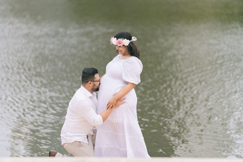 Expectant father kneels and rests his hands on his pregnant wife's belly during a maternity photo shoot by the water.