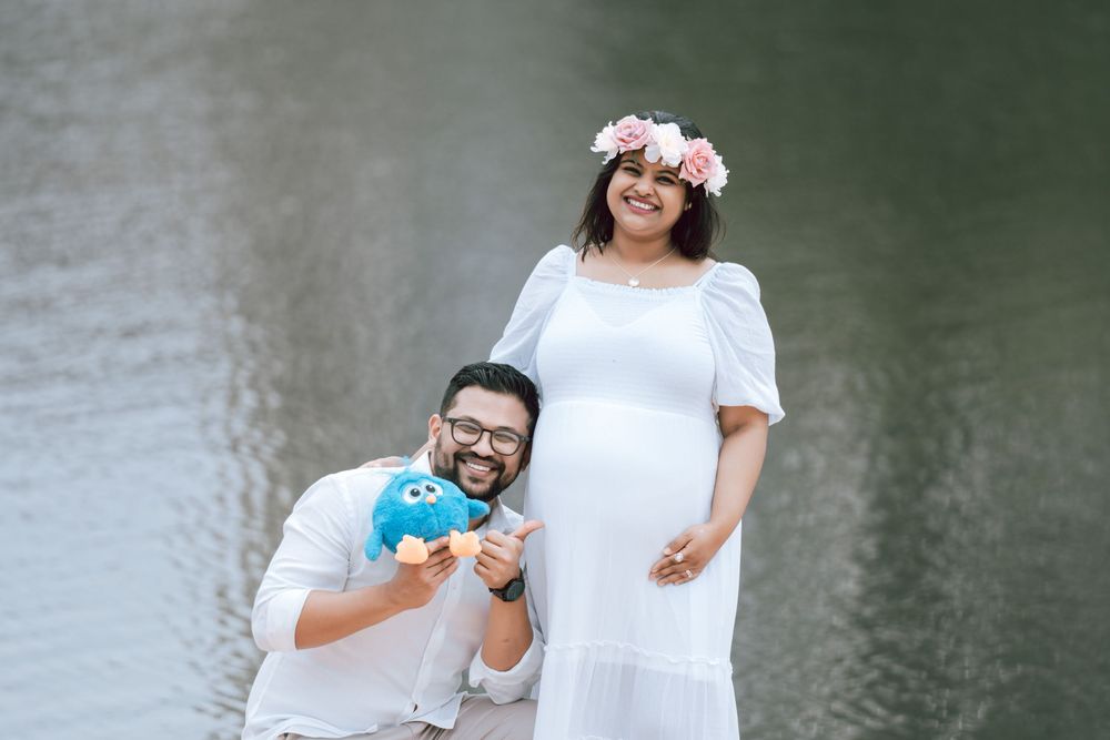 Smiling pregnant woman in a white dress and flower crown stands beside a bearded man holding a blue plush toy by a lake.