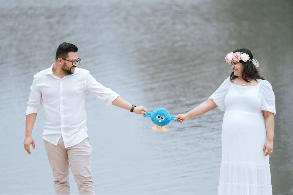 Expecting couple in white outfits holds a blue plush toy between them while standing by a calm lake, the woman wearing a floral crown.