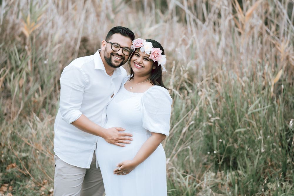 Smiling couple embracing in a maternity photo shoot, the expecting mother wearing a white dress and floral crown among tall golden grass.