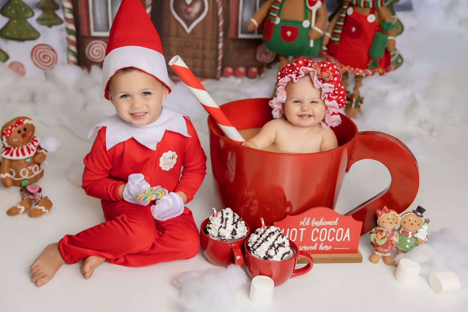 Toddler in red elf pajamas and hat sits beside a baby in a giant red hot cocoa mug in a festive Christmas gingerbread setting.