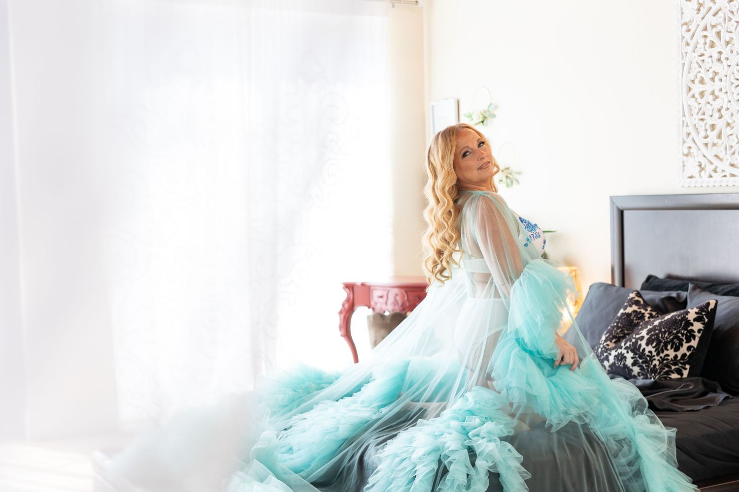 Blonde woman in a flowing turquoise tulle boudoir gown with feathered ruffles posing on a bed in a bright, airy bedroom.