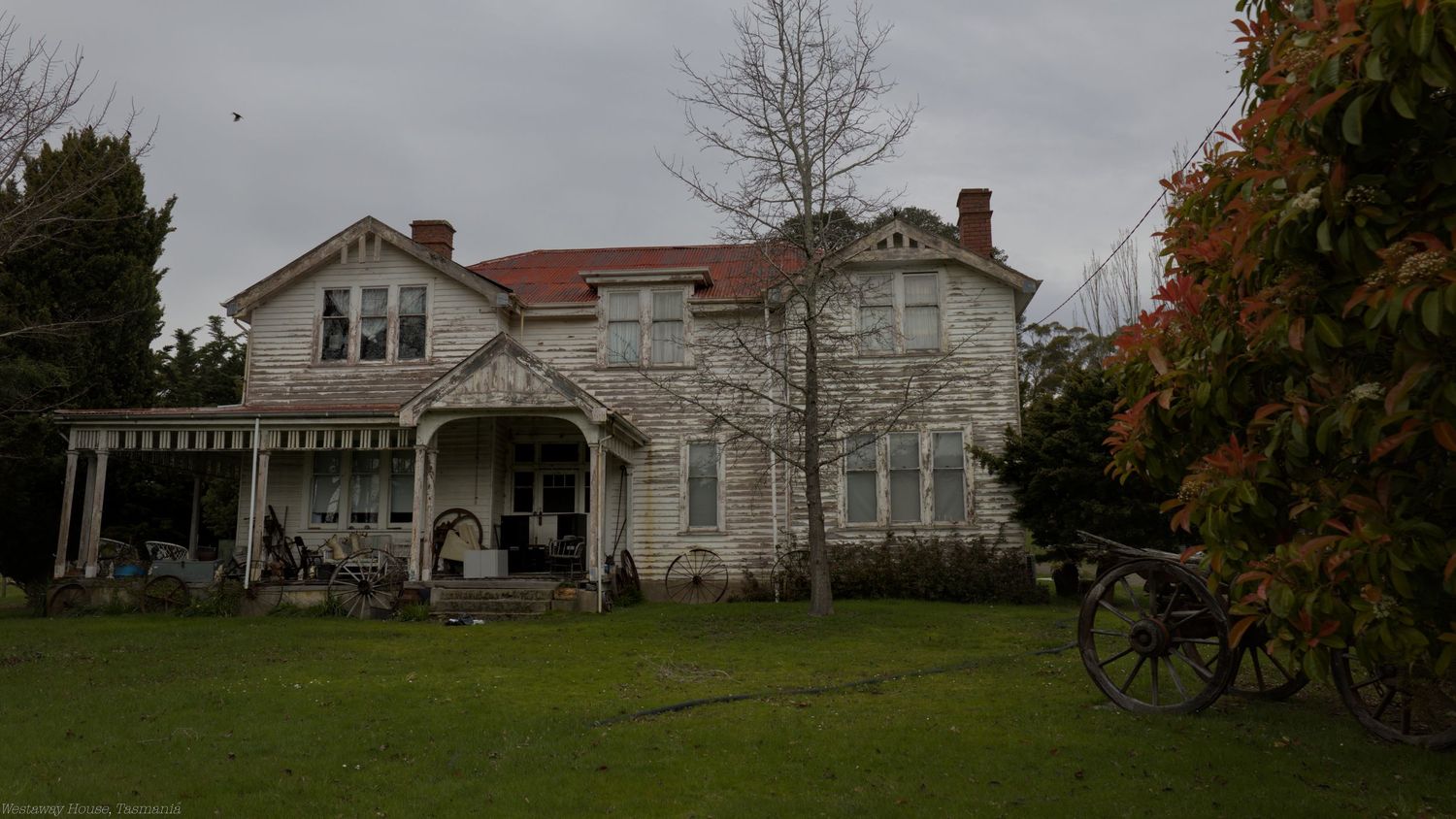 Historic two-story Victorian house with a wraparound veranda and red roof, set behind a green lawn and bare tree on an overcast day.