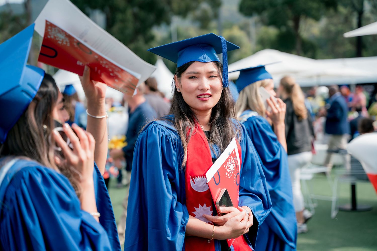 Smiling graduate in a blue cap and gown with a red stole holds a diploma folder at an outdoor graduation ceremony with fellow graduates.