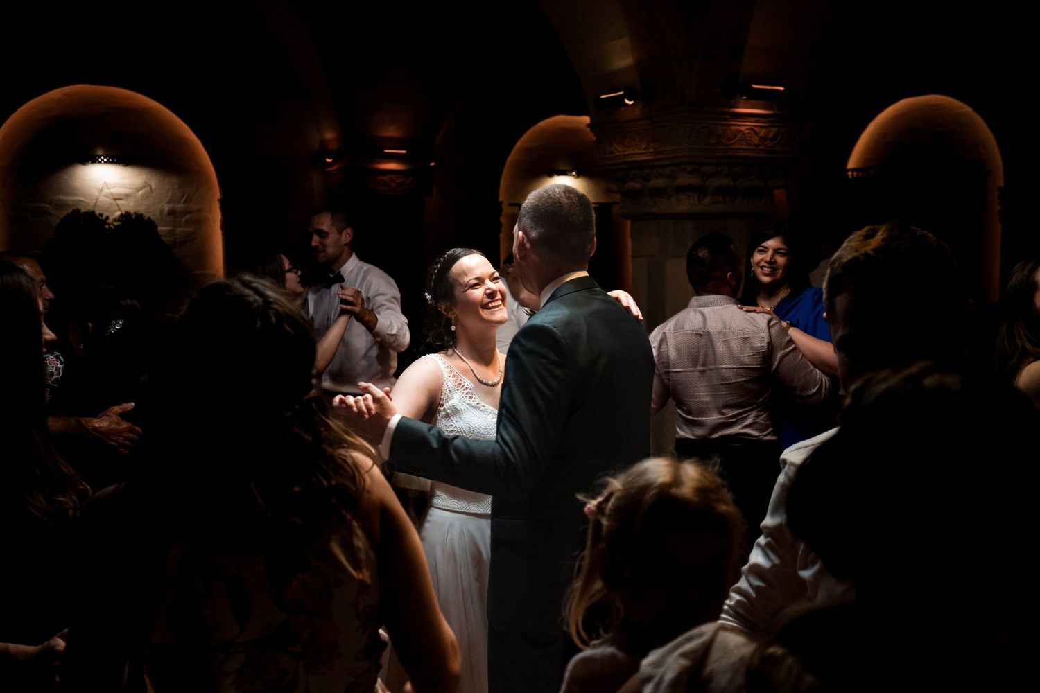 Smiling bride in a white gown dances with a groom in a dark suit as wedding guests gather around in a softly lit, arched reception hall.